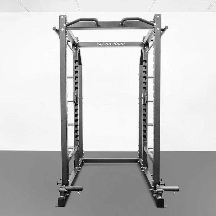 Front view of the BodyKore G703 Foundation Series Squat Cage featuring a multi-grip pull-up bar, J hooks, spotter bars, dual landmines, band pegs, and weight storage pegs, built with durable 3"x3" steel tubing for home or commercial gym use.