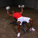 Black man in a red shirt and white shorts performing chest flys with metal hex dumbbells on a bench in an upright position.