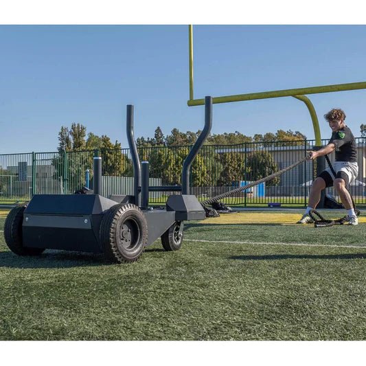  Side view of the BodyKore Smart Sled Pro on turf at an athletic field with a battle rope attached, showing a male athlete pulling the sled, demonstrating its versatility and effectiveness in outdoor strength and conditioning training.