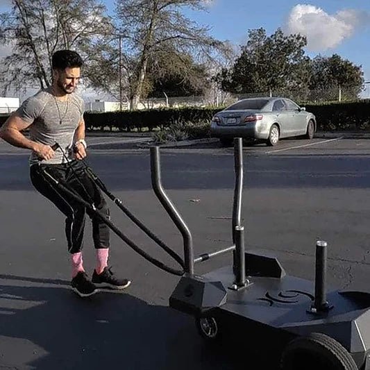 **Alt Text:** Male athlete pulling the BodyKore Smart Sled Pro on pavement, showcasing its durable all-terrain wheels and versatility for outdoor strength and conditioning workouts on various surfaces.